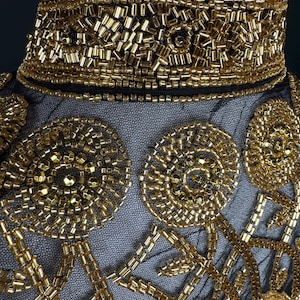 May include: Detailed view of a black mesh garment with elaborate gold bead embellishments. The design includes a high, beaded collar and circular patterns with linear details. The gold beads provide a rich, textured appearance.