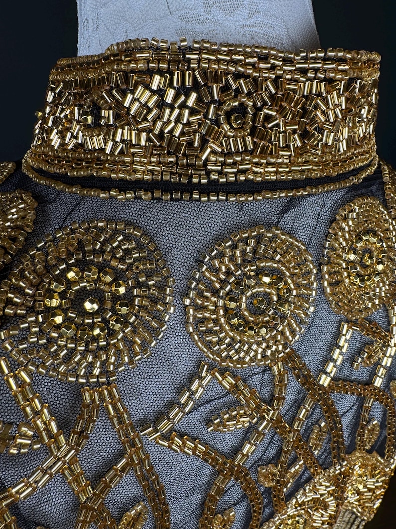 May include: Detailed view of a black mesh garment with elaborate gold bead embellishments. The design includes a high, beaded collar and circular patterns with linear details. The gold beads provide a rich, textured appearance.