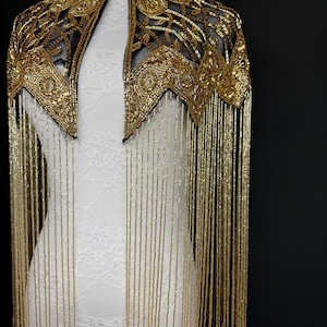 May include: A gold beaded cape with a black mesh backing. The cape features intricate floral and geometric patterns, with long, dangling gold fringe. The cape is displayed on a white mannequin against a black background.