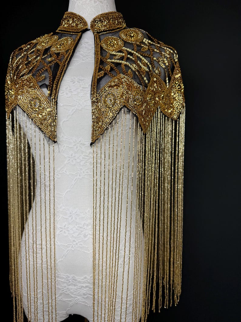 May include: A gold beaded cape with a black mesh backing. The cape features intricate floral and geometric patterns, with long, dangling gold fringe. The cape is displayed on a white mannequin against a black background.