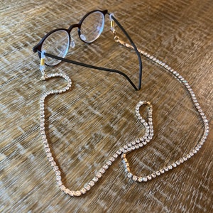 May include: A gold chain with clear rhinestones, designed to hold glasses. The chain is looped around the arms of a pair of black eyeglasses.