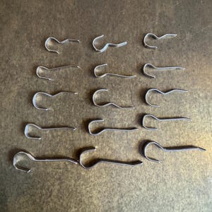May include: A collection of sixteen silver-colored cotter pins arranged on a textured, brown surface. Each pin has a looped head and a straight, pointed end, designed for securing mechanical components.