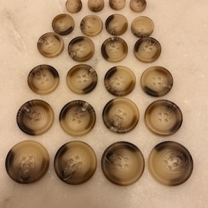 Può includere: Una collezione di bottoni marroni e beige di varie dimensioni. I bottoni hanno un aspetto marmorizzato e quattro fori per cucire. I bottoni sono disposti su una superficie chiara.