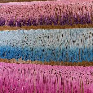 May include: A close-up of three rows of decorative fringe trim. The top row fades from pink to purple, the middle row from light blue to dark blue, and the bottom row from pink to hot pink. The fringe is made of soft, flowing threads.