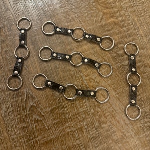Könnte beinhalten: Fünf schwarze Leder-Schlüsselanhänger mit silbernen Metallringen. Jeder Schlüsselanhänger hat mehrere Ringe, die durch ein schwarzes Lederband mit silbernen Nieten verbunden sind.