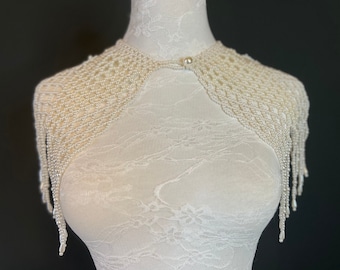 May include: Ivory-colored beaded shoulder wrap with a delicate, open-weave design. The wrap features a button closure at the neckline and long, dangling beaded fringe. The piece is displayed on a white mannequin, showcasing its elegant drape.