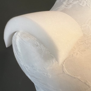 May include: White lace fabric with a textured, raised design. The fabric is draped over a form, creating a curved shape.