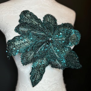 Puede incluir: Una aplicación floral de color turquesa con intrincados detalles de cuentas y lentejuelas. La flor se exhibe sobre un maniquí blanco, mostrando su delicado diseño y textura. Los pétalos están delineados con cuentas y rellenos de lentejuelas.