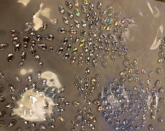 May include: Close-up of a sheet of clear adhesive-backed rhinestones. The rhinestones are iridescent and have a teardrop shape. They are arranged in a pattern of clusters.