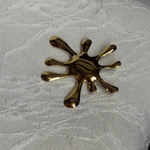 Puede incluir: Un broche abstracto de color dorado en forma de salpicadura o estallido de estrella.