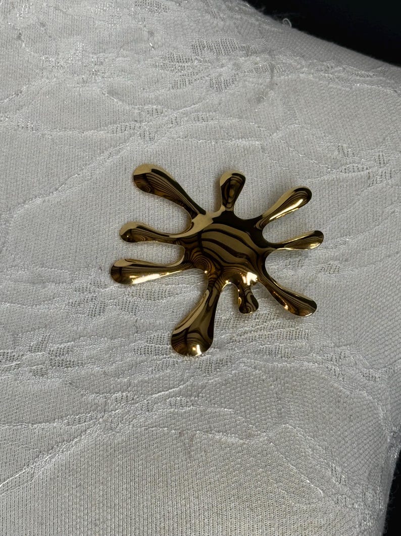Puede incluir: Un broche abstracto de color dorado en forma de salpicadura o estallido de estrella.