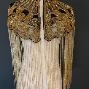 May include: A black and gold beaded cape with fringe. The cape features intricate gold beadwork on a black mesh base, with a high collar and long gold fringe cascading down. The garment is displayed on a mannequin.