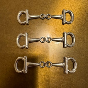 Puede incluir: Tres hebillas de cinturón de metal plateado con un diseño sencillo. Cada hebilla tiene un anillo en forma de D en cada extremo conectado por una barra recta.