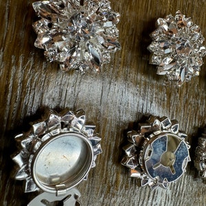 May include: Six silver-toned metal embellishments with crystal-like accents. Some of the embellishments have a circular design with a clear center, while others have a more intricate design with a raised center.