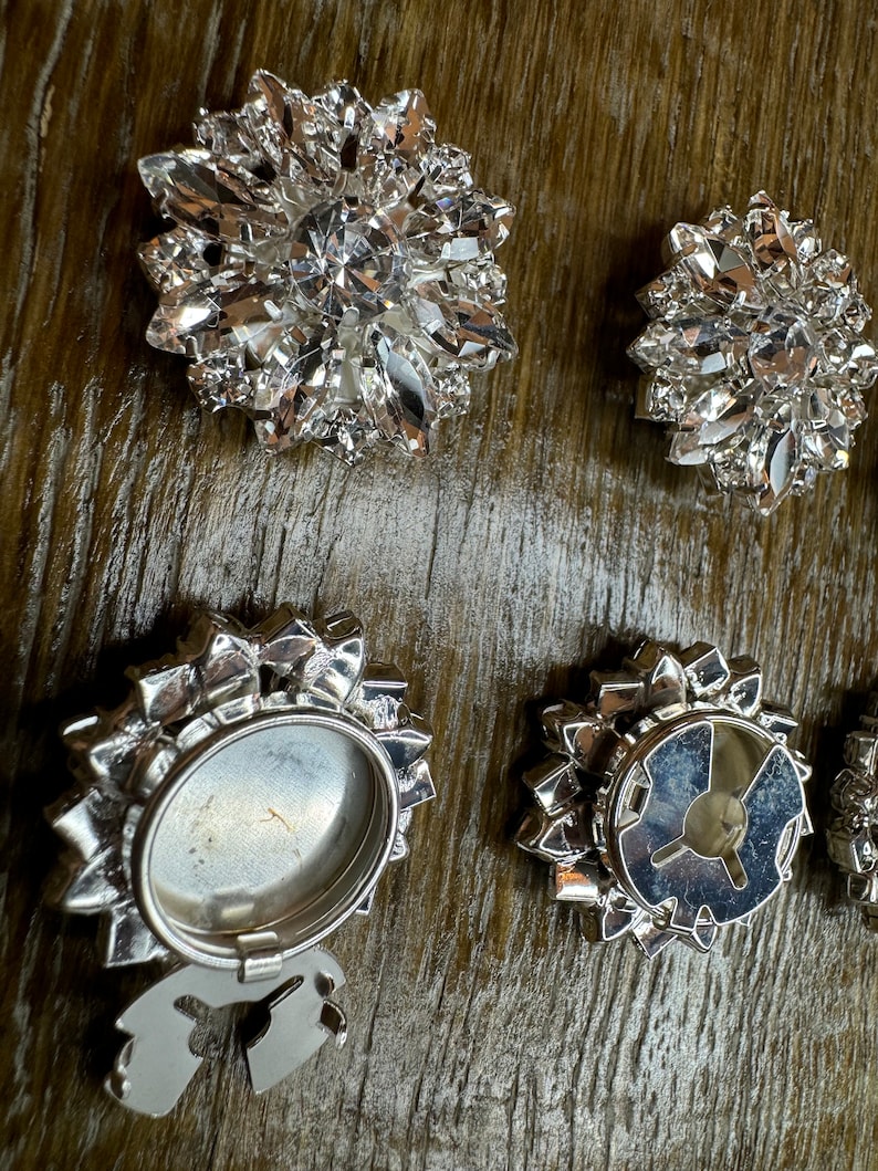 May include: Six silver-toned metal embellishments with crystal-like accents. Some of the embellishments have a circular design with a clear center, while others have a more intricate design with a raised center.
