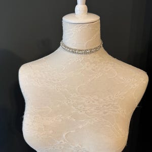 May include: A sparkling choker necklace with clear, round rhinestones set in a double-row design. The necklace is displayed on a white mannequin with a lace pattern, set against a dark background. The choker is a delicate piece of jewelry.