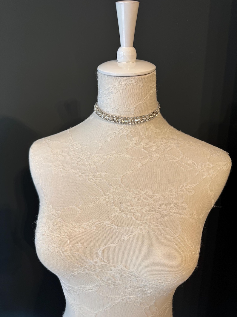 May include: A sparkling choker necklace with clear, round rhinestones set in a double-row design. The necklace is displayed on a white mannequin with a lace pattern, set against a dark background. The choker is a delicate piece of jewelry.
