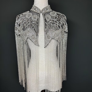 以下が含まれることがあります： ハイカラーと複雑な花柄が特徴の、シルバービーズのケープ。 ケープは、腕から垂れ下がる長いビーズフリンジが特徴です。 この衣服は、暗い灰色の背景に対して白いマネキンに展示されています。