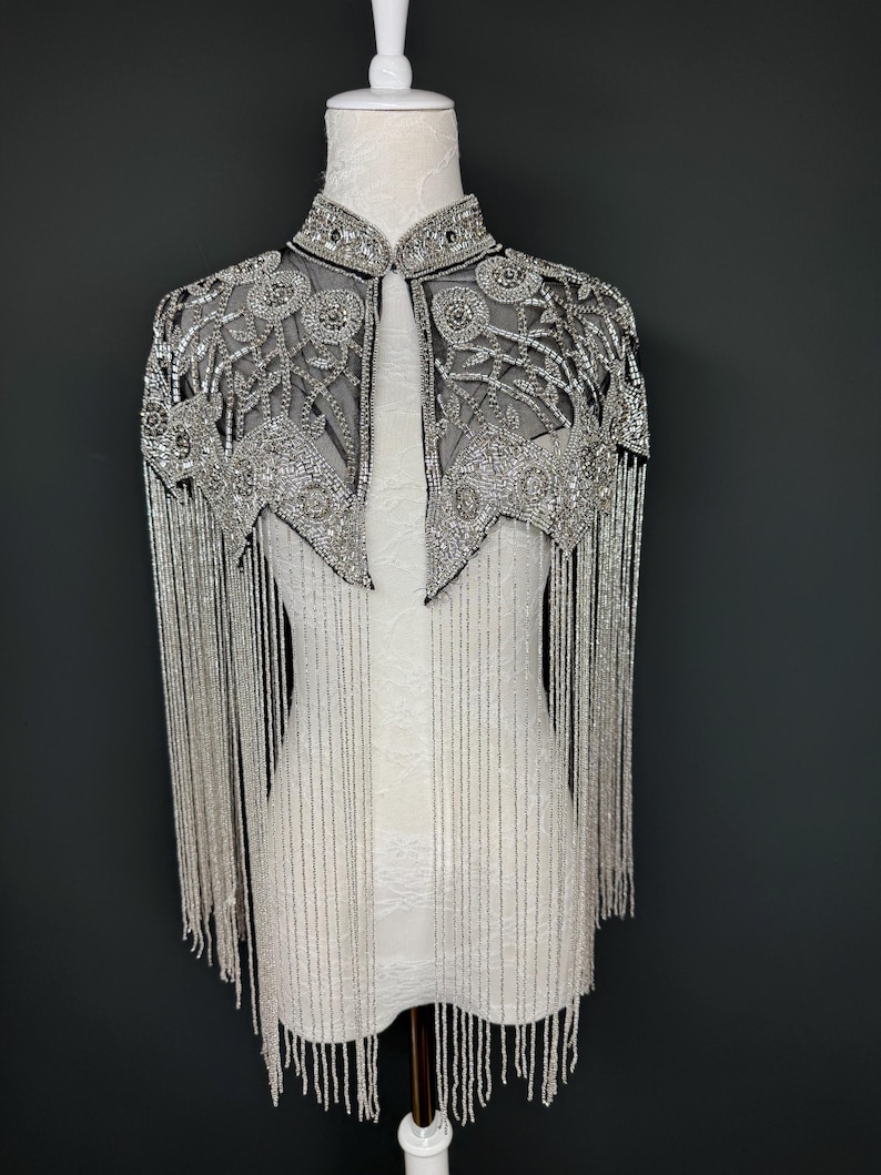 May include: A silver beaded cape with a high collar and intricate floral patterns. The cape features long beaded fringe that hangs down the arms. The garment is displayed on a white mannequin against a dark grey background.