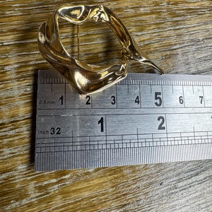 May include: A gold-coloured abstract metal brooch with an irregular, organic shape. The brooch is approximately 5 cm wide and 4 cm tall.