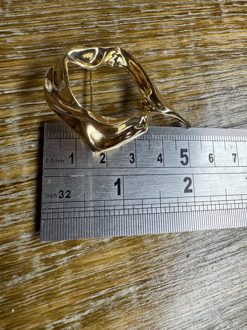 May include: A gold-coloured abstract metal brooch with an irregular, organic shape. The brooch is approximately 5 cm wide and 4 cm tall.