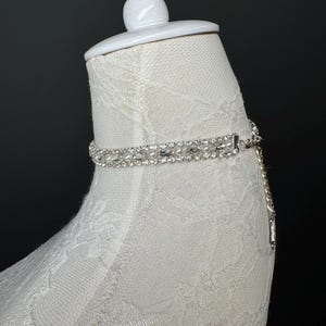 May include: A silver choker necklace with clear rhinestones and small white pearls. The necklace is displayed on a white mannequin head covered in white lace. A silver chain hangs from the clasp.