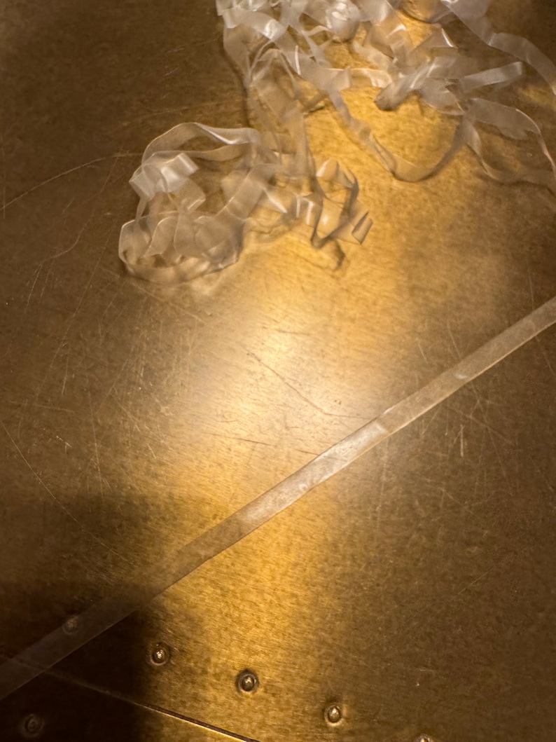 May include: A close-up of a gold surface with a clear plastic tape strip and a pile of clear plastic tape. The surface has a textured pattern and several small, round, gold metal accents.