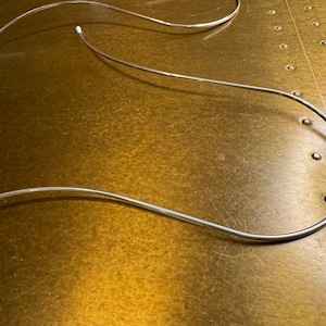 Op de afbeelding: Twee zilveren metalen buizen met witte kunststof uiteinden. De buizen zijn gebogen in een gebogen vorm en liggen op een bruin oppervlak.