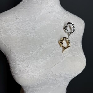 May include: Two abstract metal brooches, one gold and one silver, are pinned to a white fabric mannequin. The brooches are both in the shape of a twisted oval.