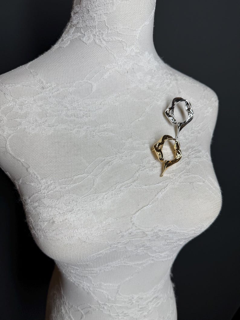 May include: Two abstract metal brooches, one gold and one silver, are pinned to a white fabric mannequin. The brooches are both in the shape of a twisted oval.