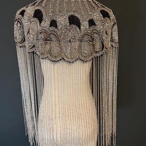 May include: A beaded cape with a high collar and long fringe. The cape features intricate silver and black beadwork in a decorative pattern. The fringe hangs down from the cape's edge. The cape is displayed on a mannequin.