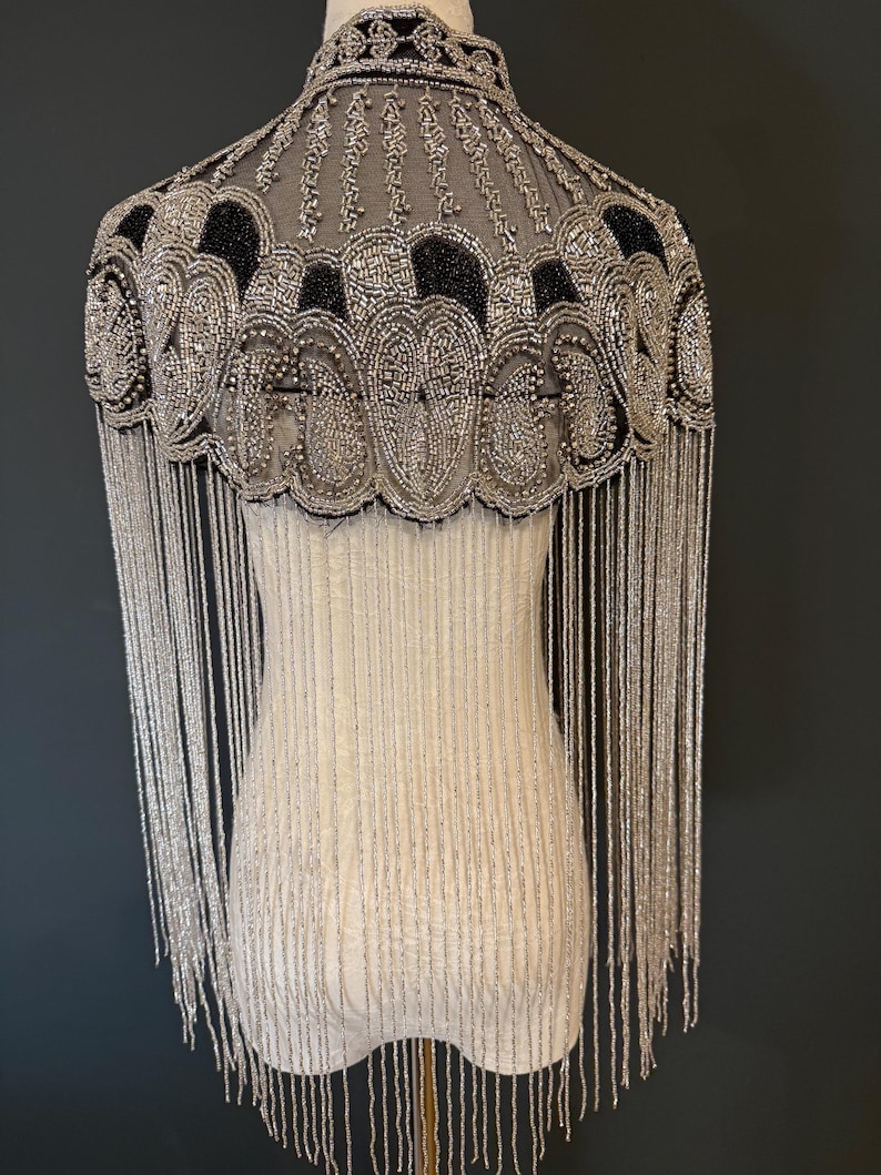 May include: A beaded cape with a high collar and long fringe. The cape features intricate silver and black beadwork in a decorative pattern. The fringe hangs down from the cape's edge. The cape is displayed on a mannequin.