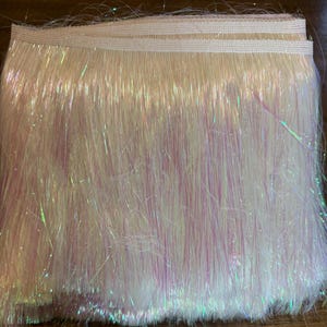 Puede incluir: Un paquete de flecos iridiscentes en tonos blanco, rosa y verde. Los flecos están unidos a una banda elástica de color crema. El material tiene un aspecto brillante y texturizado, con hebras de diferentes longitudes.