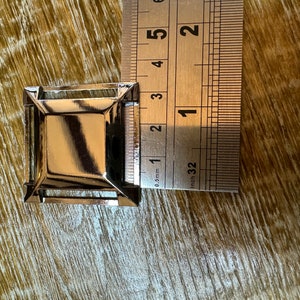 Op de afbeelding: Een vierkante gesp van zilverkleurig metaal met een verhoogd midden, ongeveer 1 inch groot.