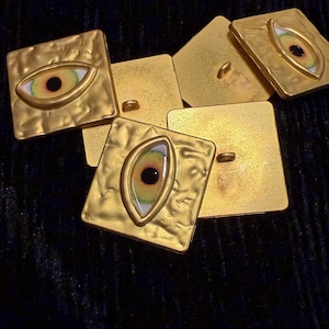May include: Several gold-coloured, square buttons with a textured surface. Each button features a painted eye design with a black pupil, surrounded by yellow, green, and white. The buttons are arranged on a dark background.