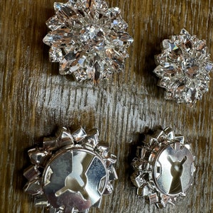 May include: Four silver-toned metal embellishments with clear crystal accents. Two of the embellishments have a circular design with a center hole. The other two embellishments have a more intricate design with a central crystal.