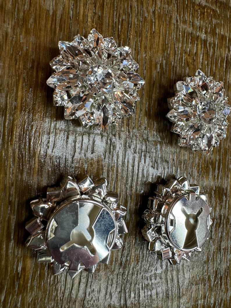 May include: Four silver-toned metal embellishments with clear crystal accents. Two of the embellishments have a circular design with a center hole. The other two embellishments have a more intricate design with a central crystal.