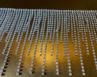 May include: A close-up of a silver fringe trim made of small, clear rhinestones. The fringe is hanging down in rows, creating a shimmering effect.