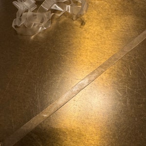 May include: A close-up of a gold surface with two pieces of clear plastic ribbon. The ribbon is lying on the surface and is slightly overlapping. The surface has a textured, worn look.