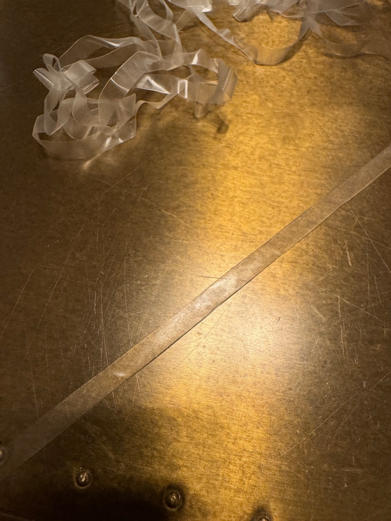 May include: A close-up of a gold surface with two pieces of clear plastic ribbon. The ribbon is lying on the surface and is slightly overlapping. The surface has a textured, worn look.