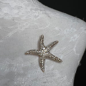 May include: A silver starfish brooch with clear rhinestones. The brooch is on a white fabric background.