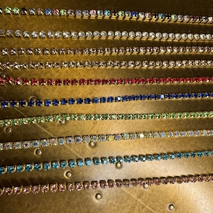 Puede incluir: Primer plano de diez filas de cadena dorada con piedras de cristal de diferentes colores. Las piedras de cristal son de varios colores, incluyendo transparente, azul claro, verde, rojo y azul.