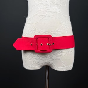Peut inclure: Une large ceinture en tissu rouge vif dotée d'une grande boucle carrée assortie. La ceinture présente une extrémité pointue et des œillets métalliques argentés pour le réglage, présentée sur un mannequin en dentelle blanche sur fond sombre.