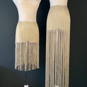 May include: Two mannequins display gold fringe skirts. The skirts are attached to a white bodice and hang to different lengths. The fringe is a shimmering gold colour. The mannequins stand against a dark background.