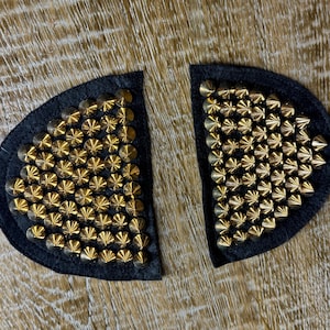 May include: Two black fabric patches with gold-coloured studs. The patches are shaped like half circles and are covered in small, evenly spaced studs.