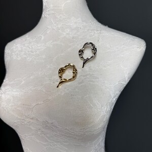 May include: Two abstract metal rings, one gold and one silver, are displayed on a white mannequin. The rings are made of twisted metal and have an organic, flowing shape.