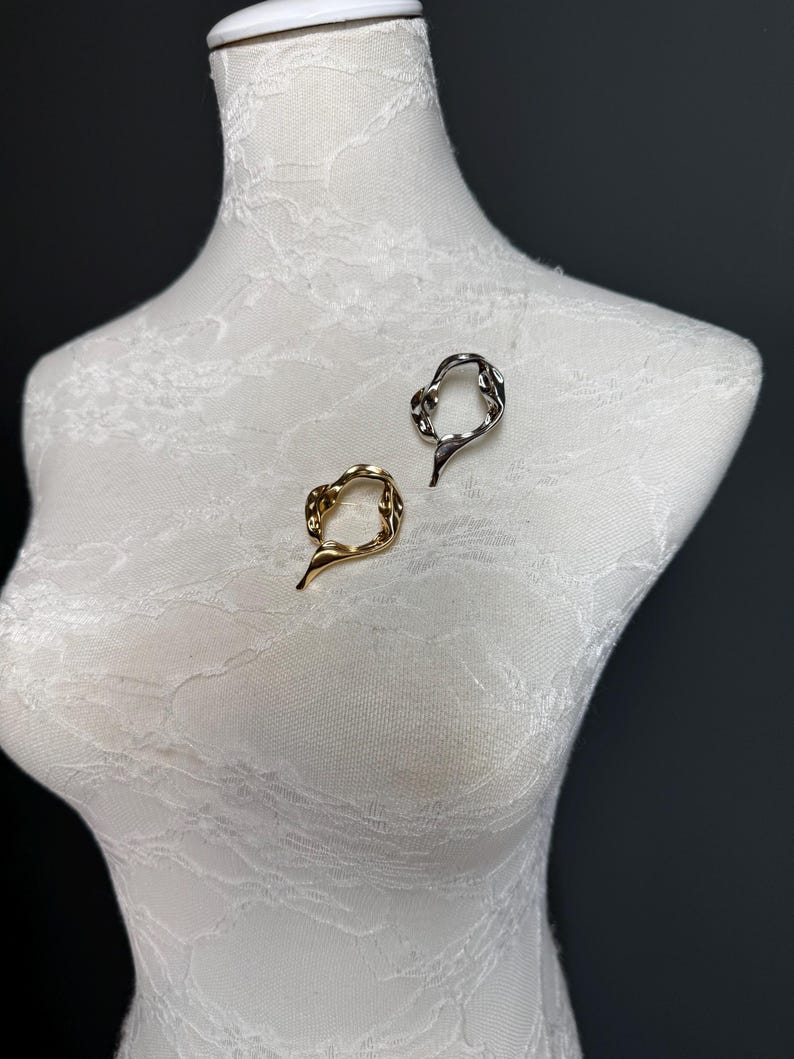 May include: Two abstract metal rings, one gold and one silver, are displayed on a white mannequin. The rings are made of twisted metal and have an organic, flowing shape.