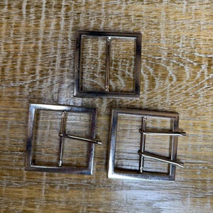 May include: Three square, silver-coloured belt buckles. Each buckle has a rectangular frame with a single bar across the centre and a pin for securing a belt. The buckles are displayed on a light brown wooden surface.