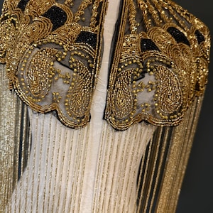 May include: A gold beaded cape with black accents and fringe. The cape features intricate patterns and a high collar. The gold beads are sewn onto a sheer black fabric, creating a luxurious and ornate design. The cape is a statement piece.