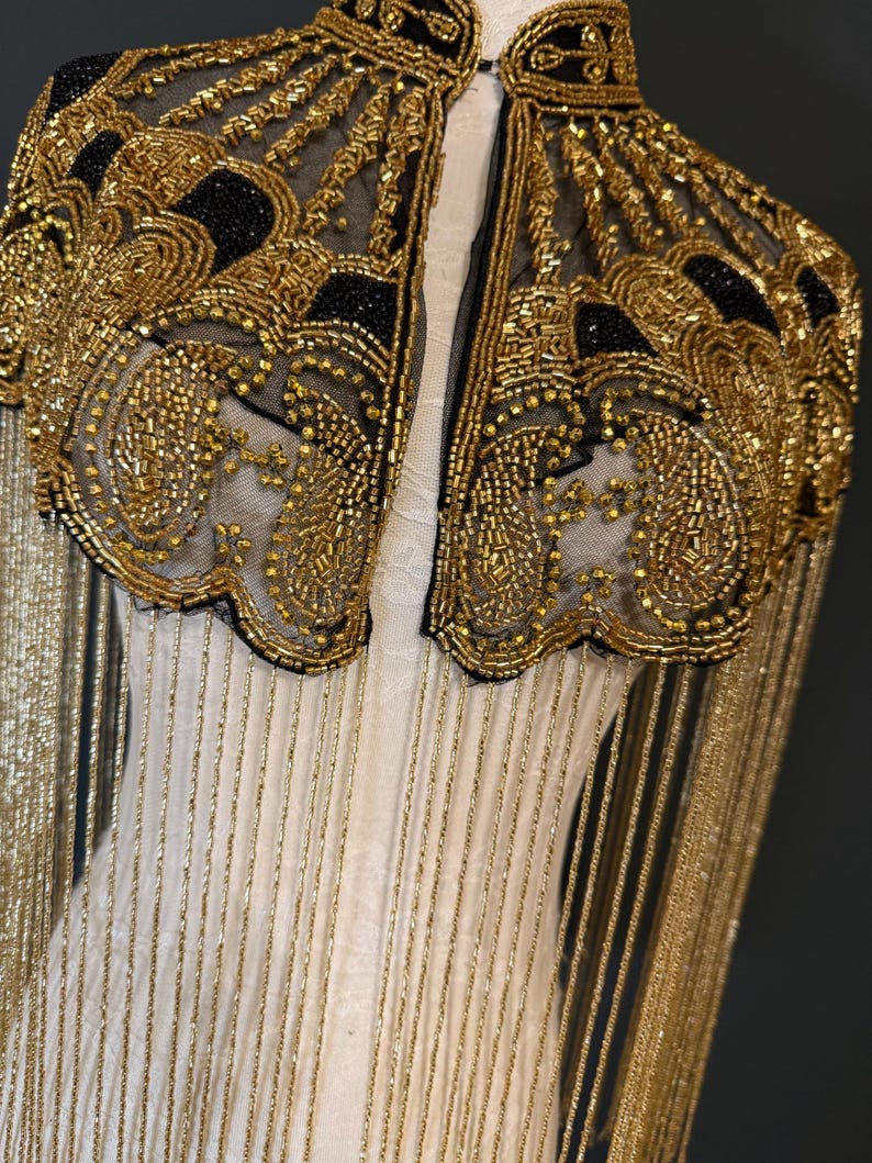 May include: A gold beaded cape with black accents and fringe. The cape features intricate patterns and a high collar. The gold beads are sewn onto a sheer black fabric, creating a luxurious and ornate design. The cape is a statement piece.